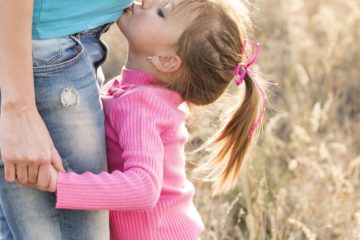 girl in pink sweater and grey jeans kissing tummy of pregnant woman in blue shirt and blue denim jeans