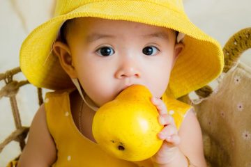 toddler in yellow top and hat holding fruit