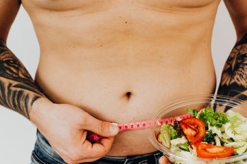 shirtless man holding a measuring tape and a bowl of vegetables